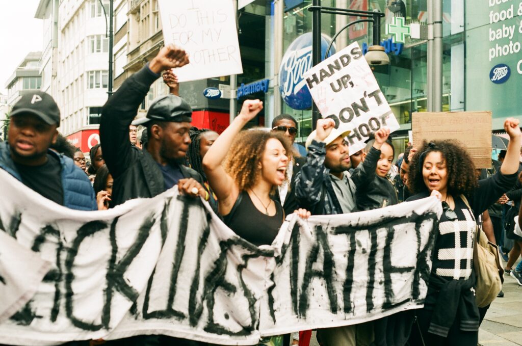 Group of 5 young black people holding a home-made banner which says "#BLACKLIVESMATTER", they are joined by a large crowd in a high street in Central London